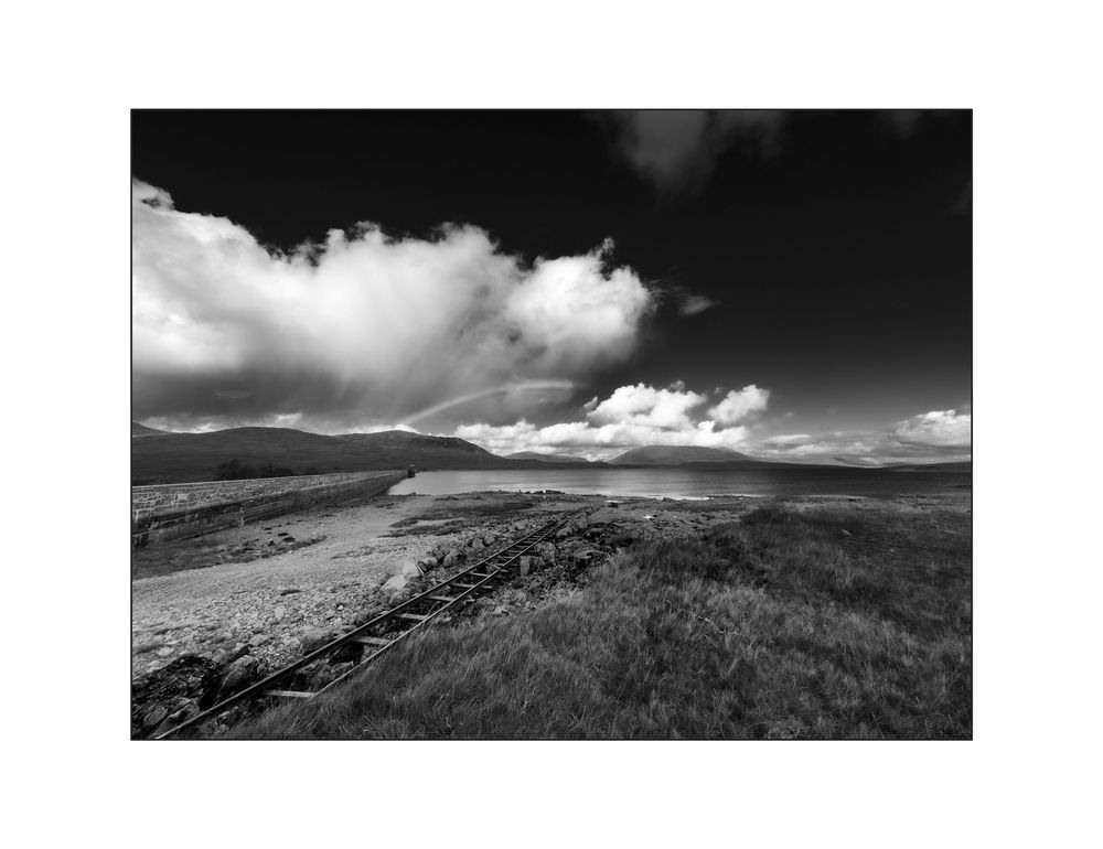 Black and white landscape photo of vast moorland with dramatic clouds, rolling hills, and wooden fence posts receding into distance.