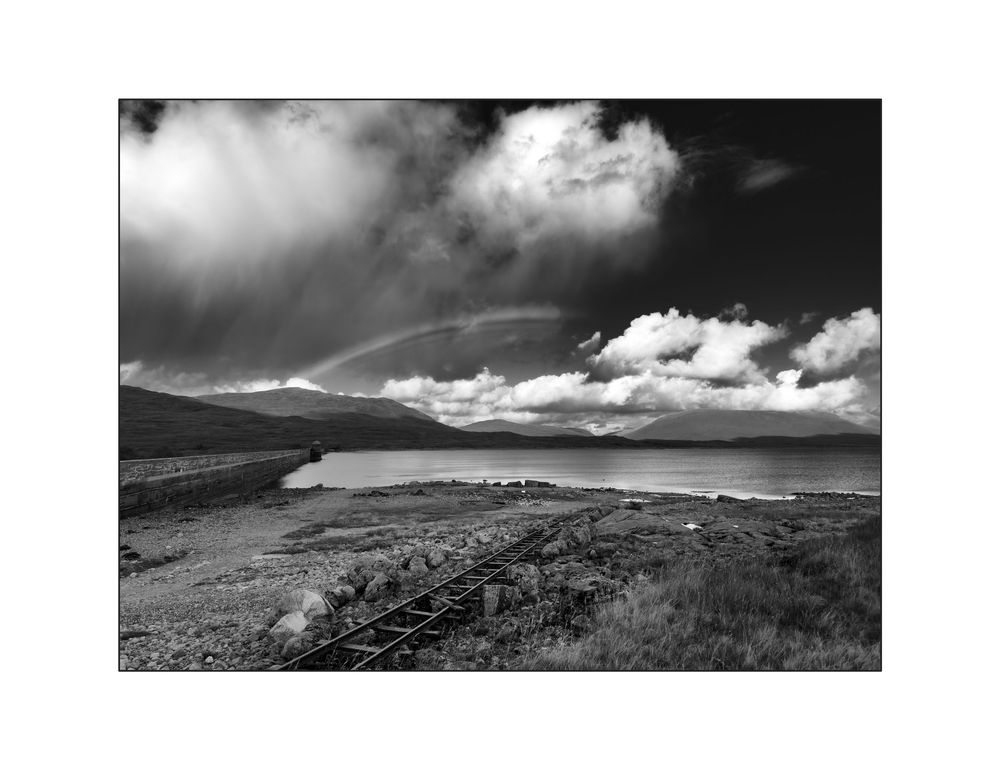 Black and white landscape photo of railway tracks leading to a loch with a rainbow and dramatic clouds in the Scottish Highlands.