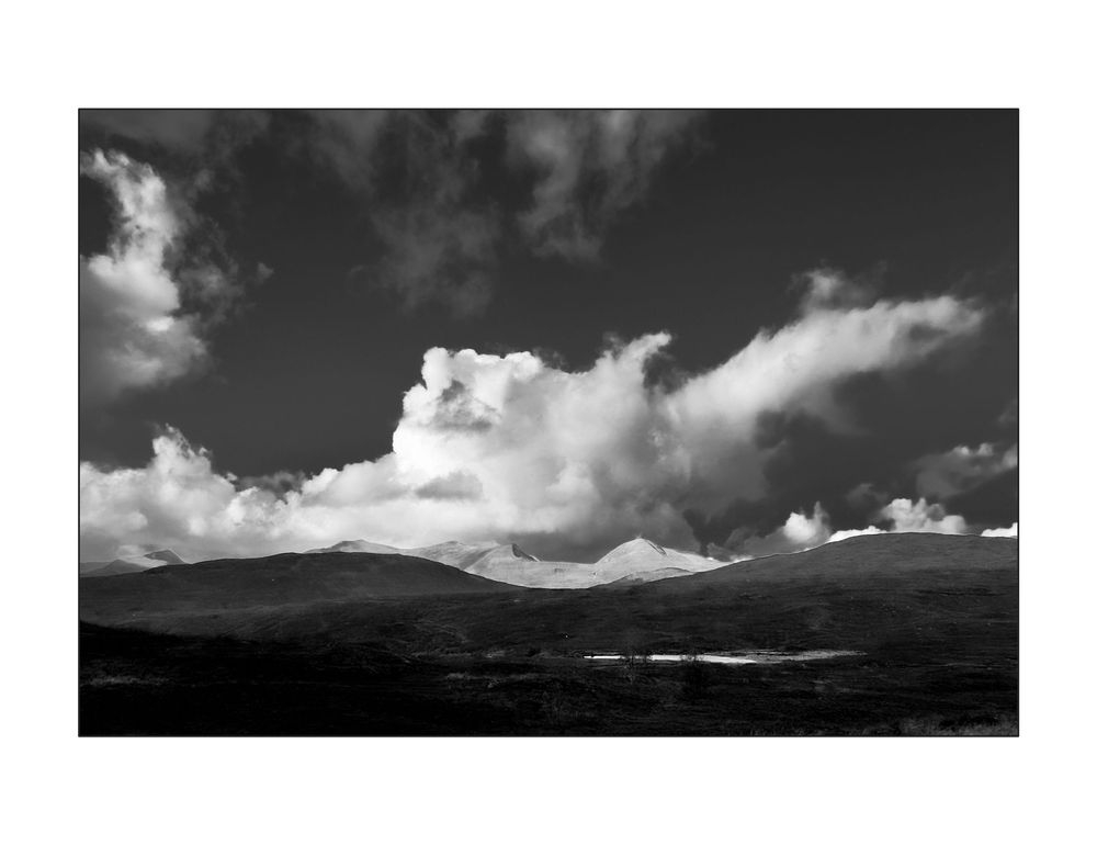 Black and white landscape photo of mountain peaks under dramatic clouds in a moody sky.