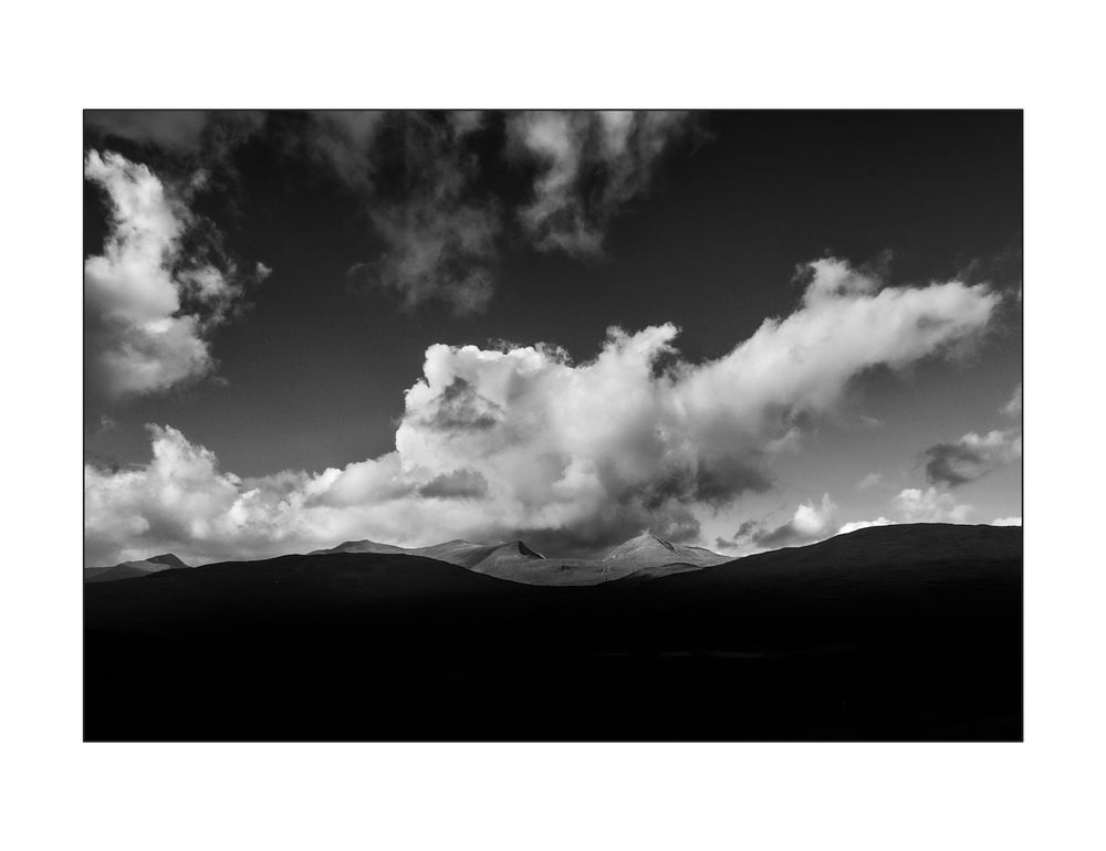 Black and white landscape photo of rolling hills silhouetted against a dramatic cloudy sky.