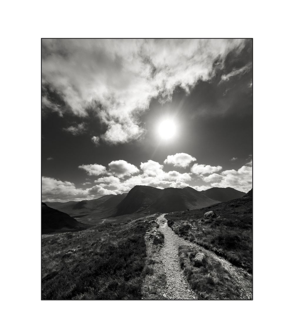 Black and white landscape photo of a mountain path leading toward peaks under a dramatic sunlit cloudy sky.