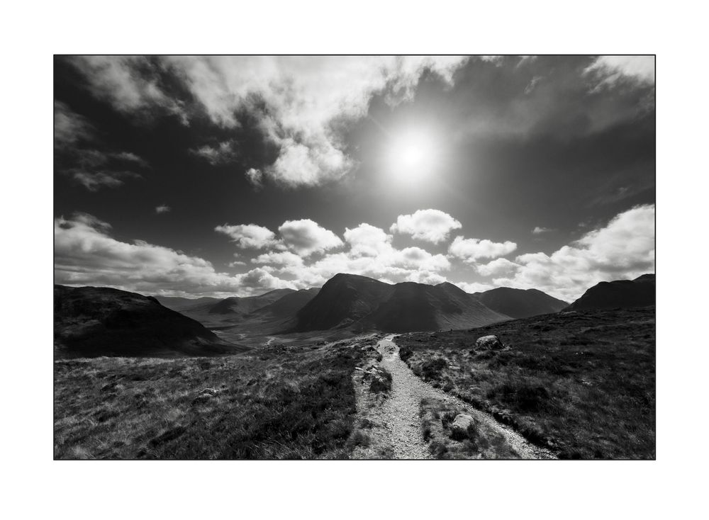 Black and white landscape photo of a rocky mountain path leading through moorland under a dramatic sunlit sky.