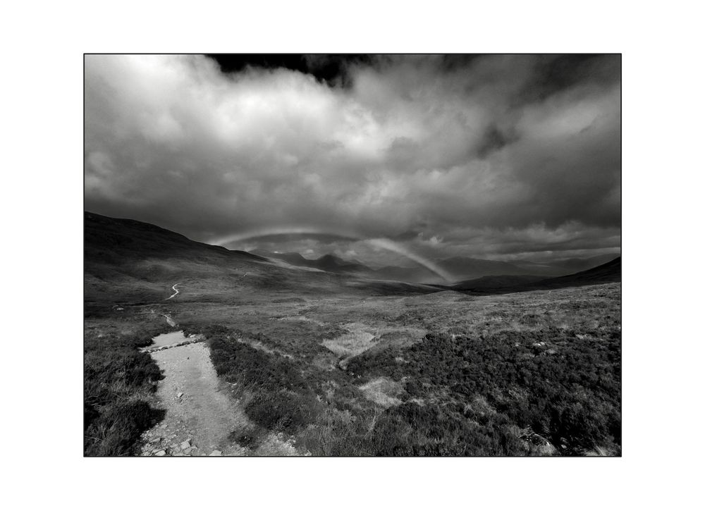 Black and white landscape photo of Scottish highlands with dramatic storm clouds and faint rainbow over rolling hills.