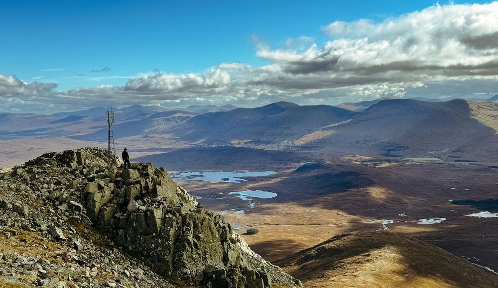 A panoramic mountain landscape view with dramatic clouds, rocky peaks, and distant lakes stretching across the horizon.