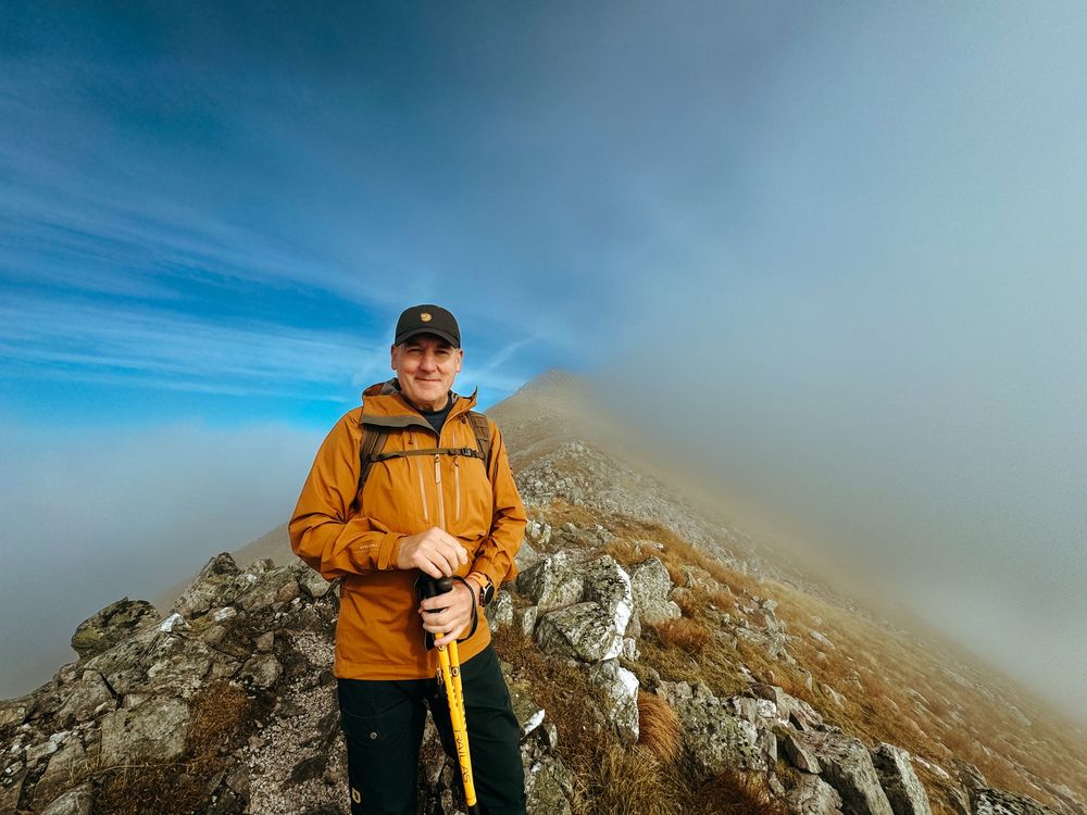 Hiker in yellow jacket stands on rocky mountain ridge with dramatic misty clouds and blue sky in background.