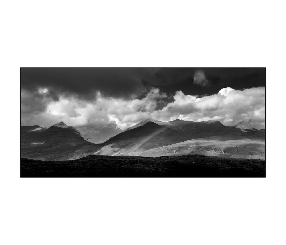 Black and white panoramic landscape of dramatic mountain peaks under stormy clouds with rays of light breaking through.