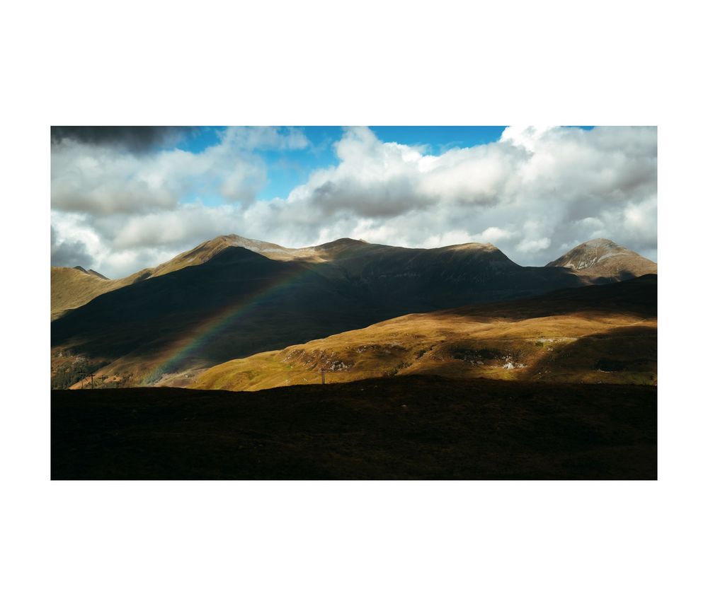 A vibrant rainbow arches over dramatic mountain peaks under a cloudy sky in a stunning highland landscape.