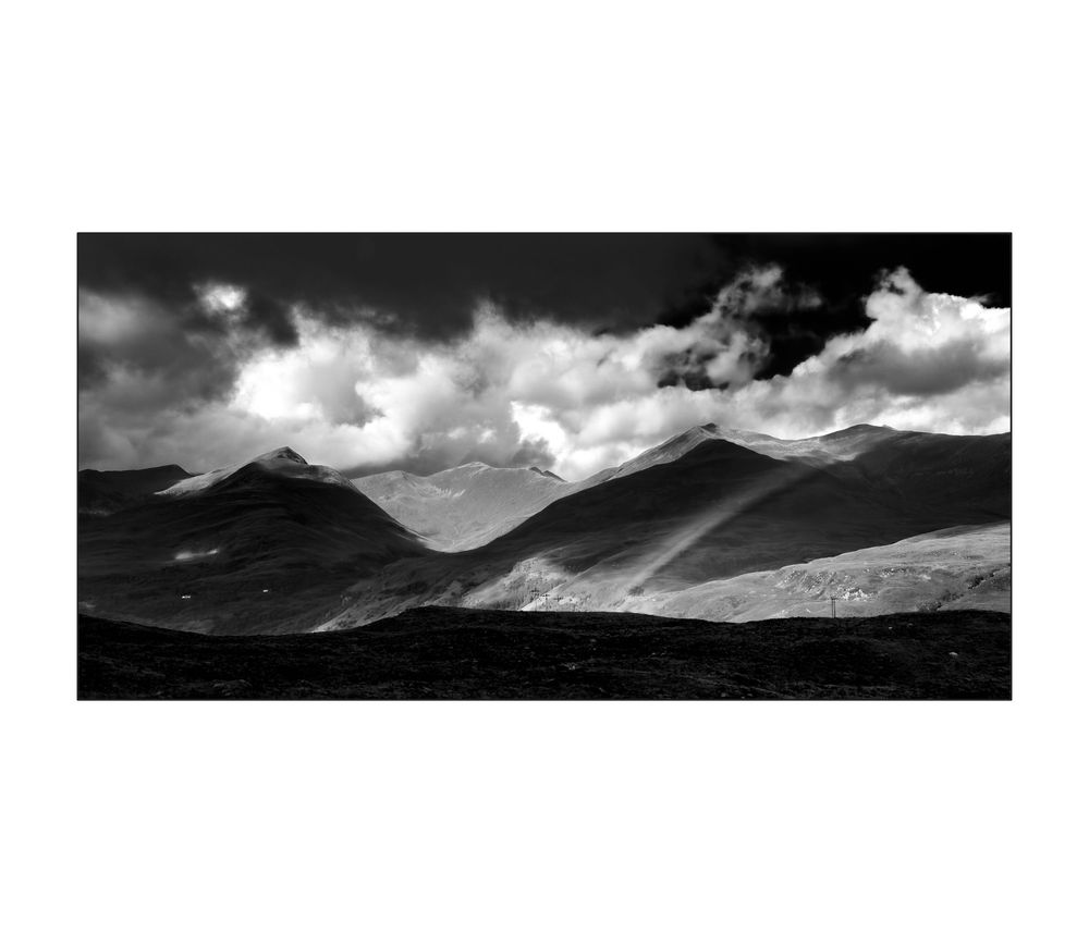 Black and white landscape photo of dramatic mountain peaks under billowing clouds with rays of light.