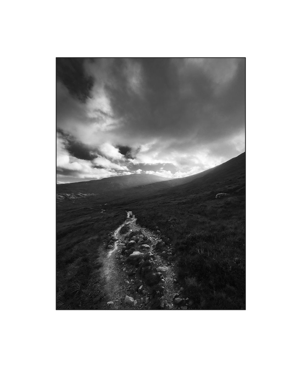 Black and white landscape photo of a narrow path through moorland under dramatic cloudy sky.