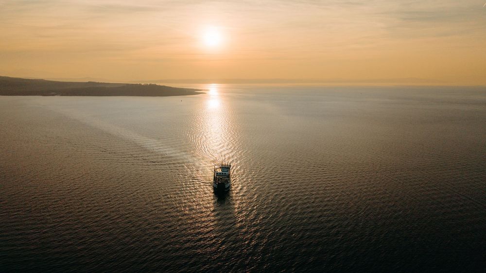 A sailboat silhouetted against a golden sunset on calm waters with distant shoreline visible on the horizon.