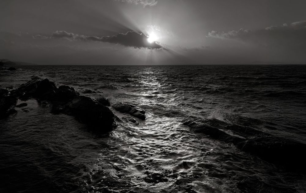 Dramatic black and white ocean sunset with sunlight reflecting off rippling waves and rocky coastline silhouettes.