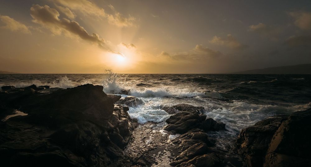 Dramatic panoramic seascape showing stormy ocean waves crashing against rocky cliffs at sunset with golden clouds.