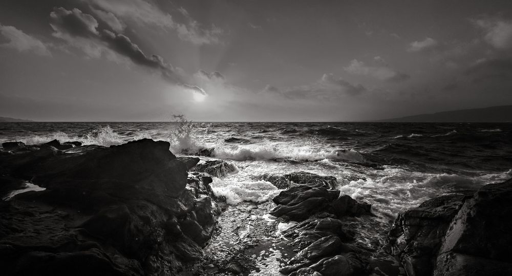 Dramatic black and white seascape with crashing waves against rocky shore during sunset over turbulent ocean.