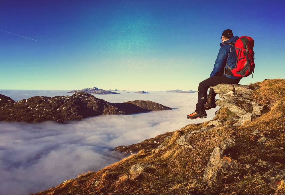Hiker with red backpack sits on rocky mountain peak above misty clouds during sunset.