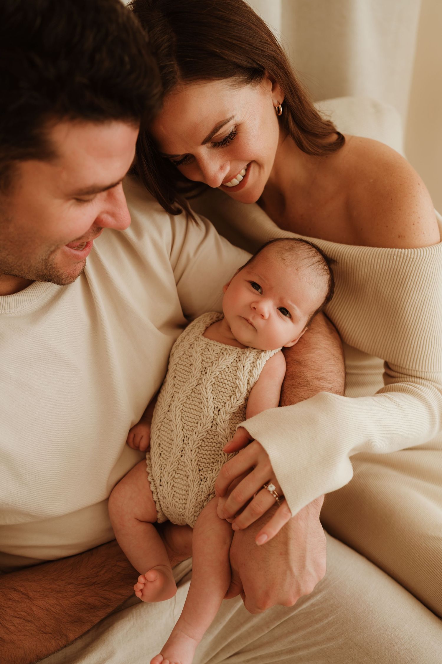 A loving family moment captured in warm natural light as parents embrace their smiling baby in matching white outfits.