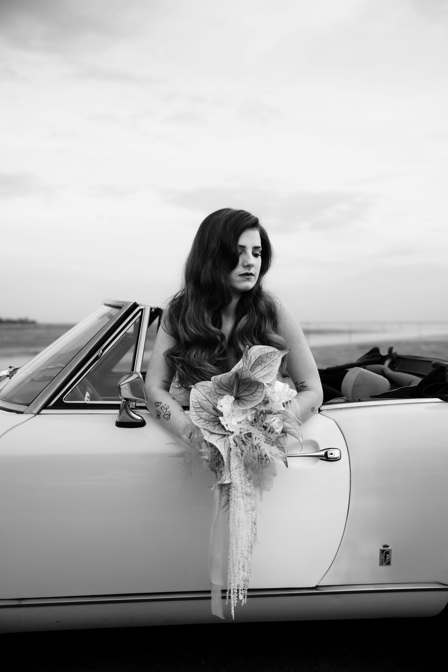A bride poses elegantly against a vintage convertible car in a black and white portrait.