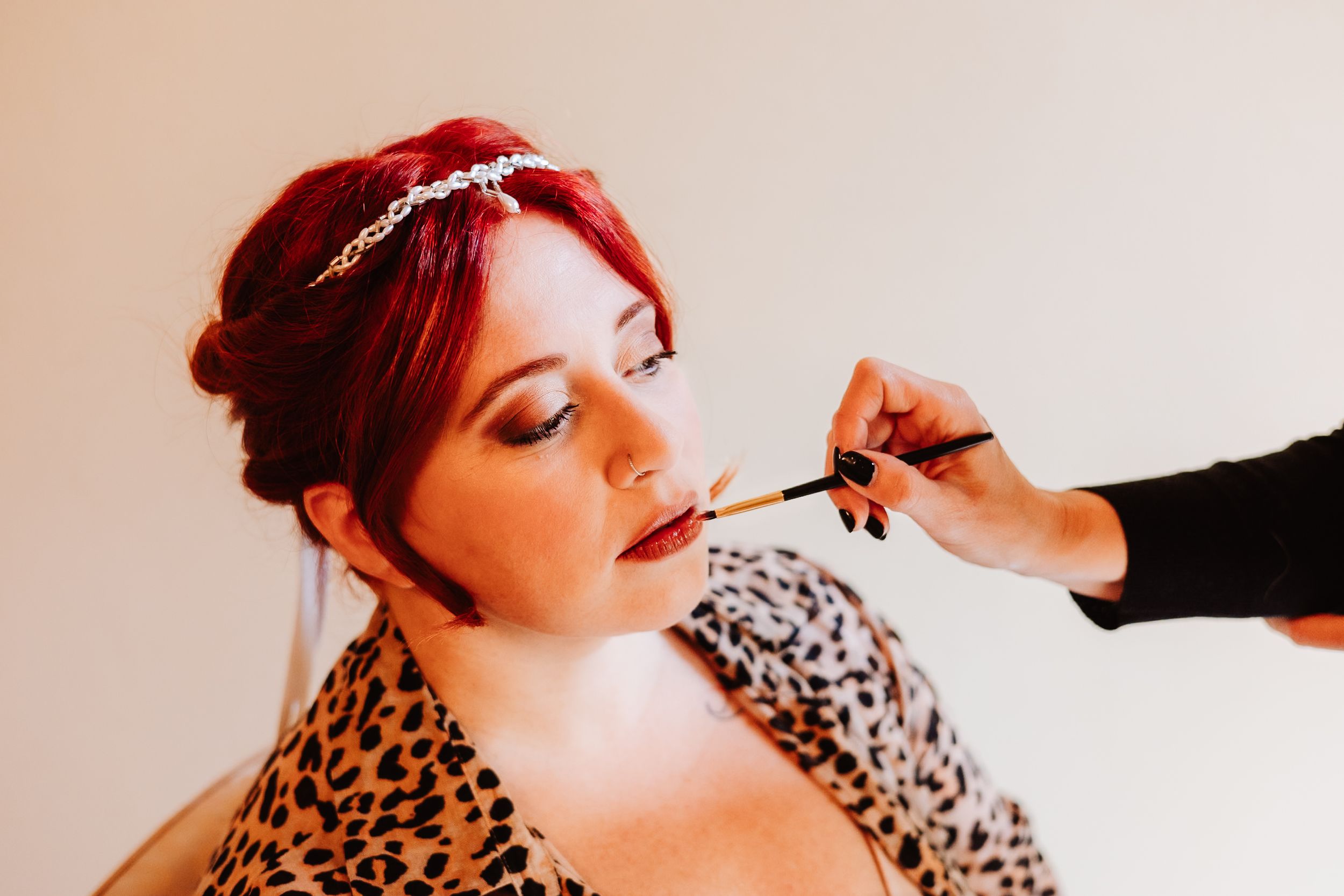 A makeup artist applies lipstick to someone wearing an animal print top and sparkly hair accessory.