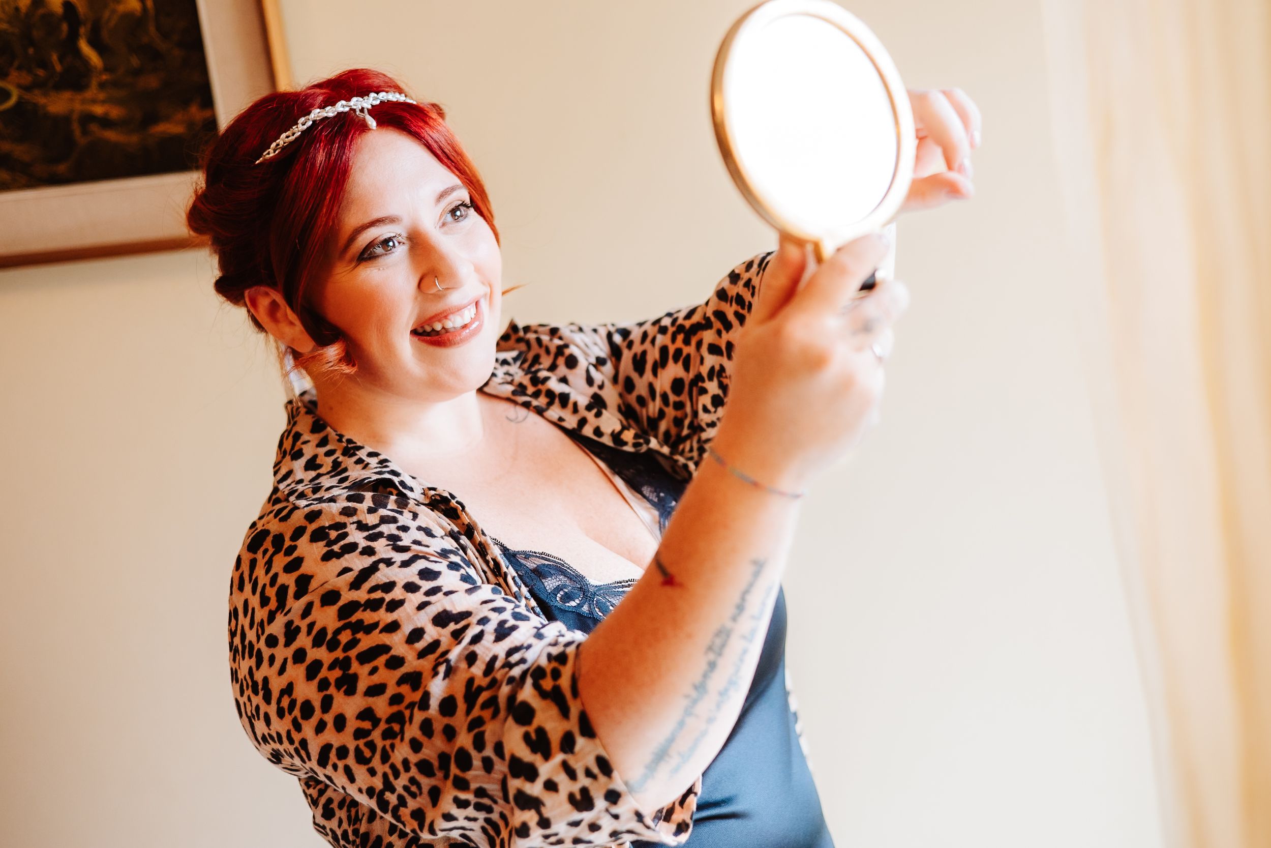 Person in a leopard print top and red headband holding a mirror while posing against a wall.