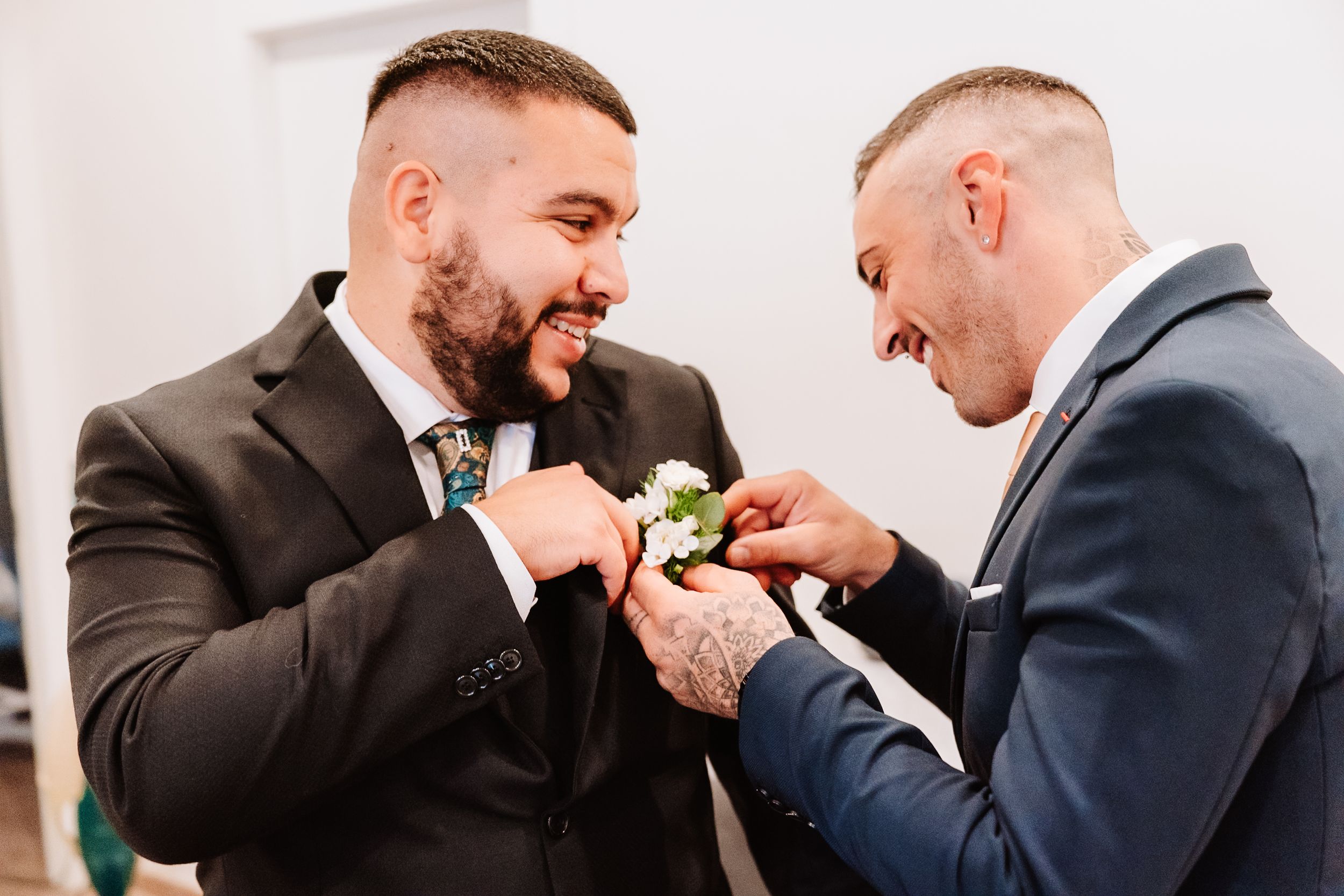 Two people in dark suits share a lighthearted moment while adjusting boutonniere flowers during a formal event.