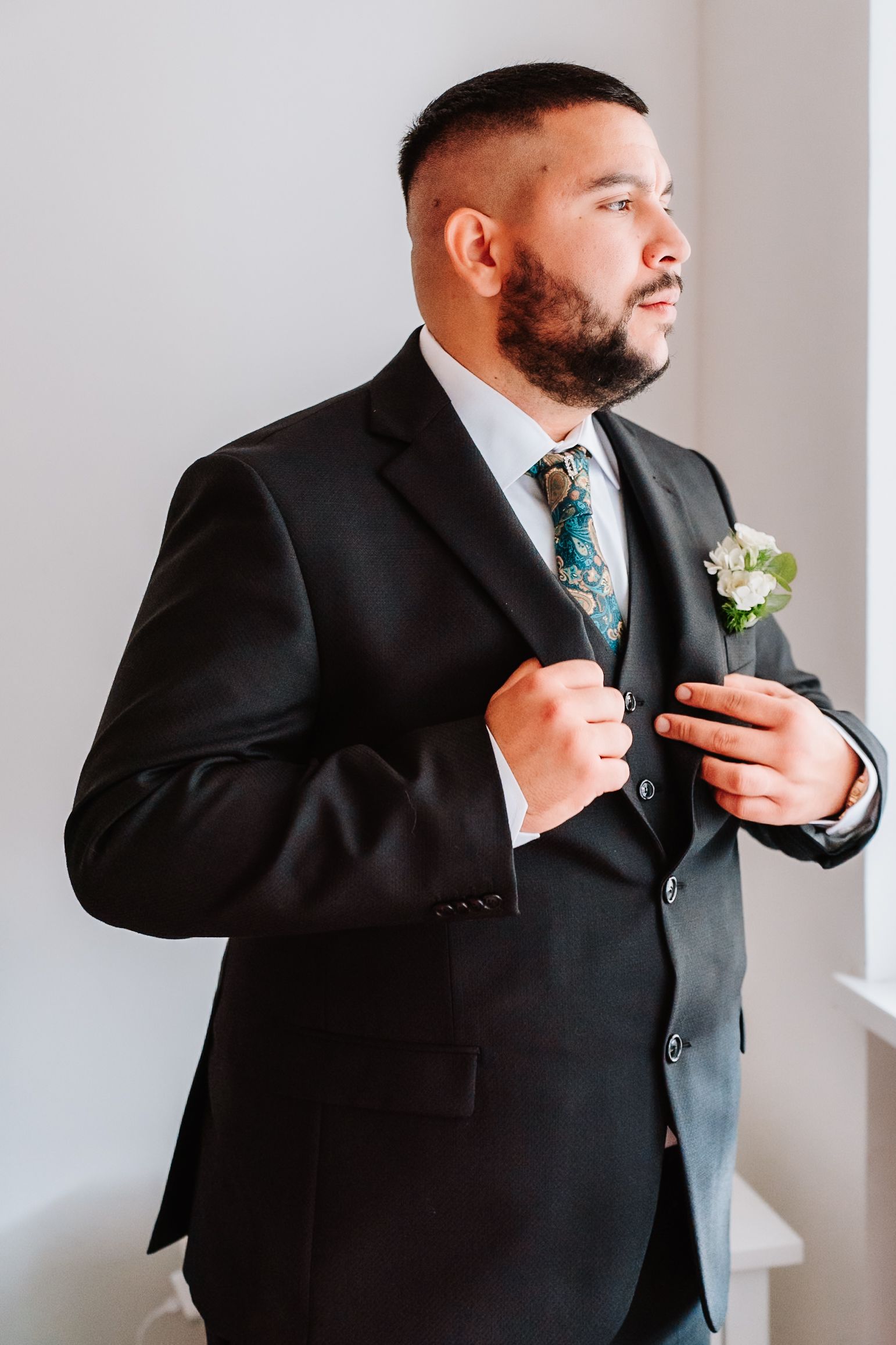 Professional groom in black suit with teal patterned tie adjusts his boutonniere while getting ready for wedding.