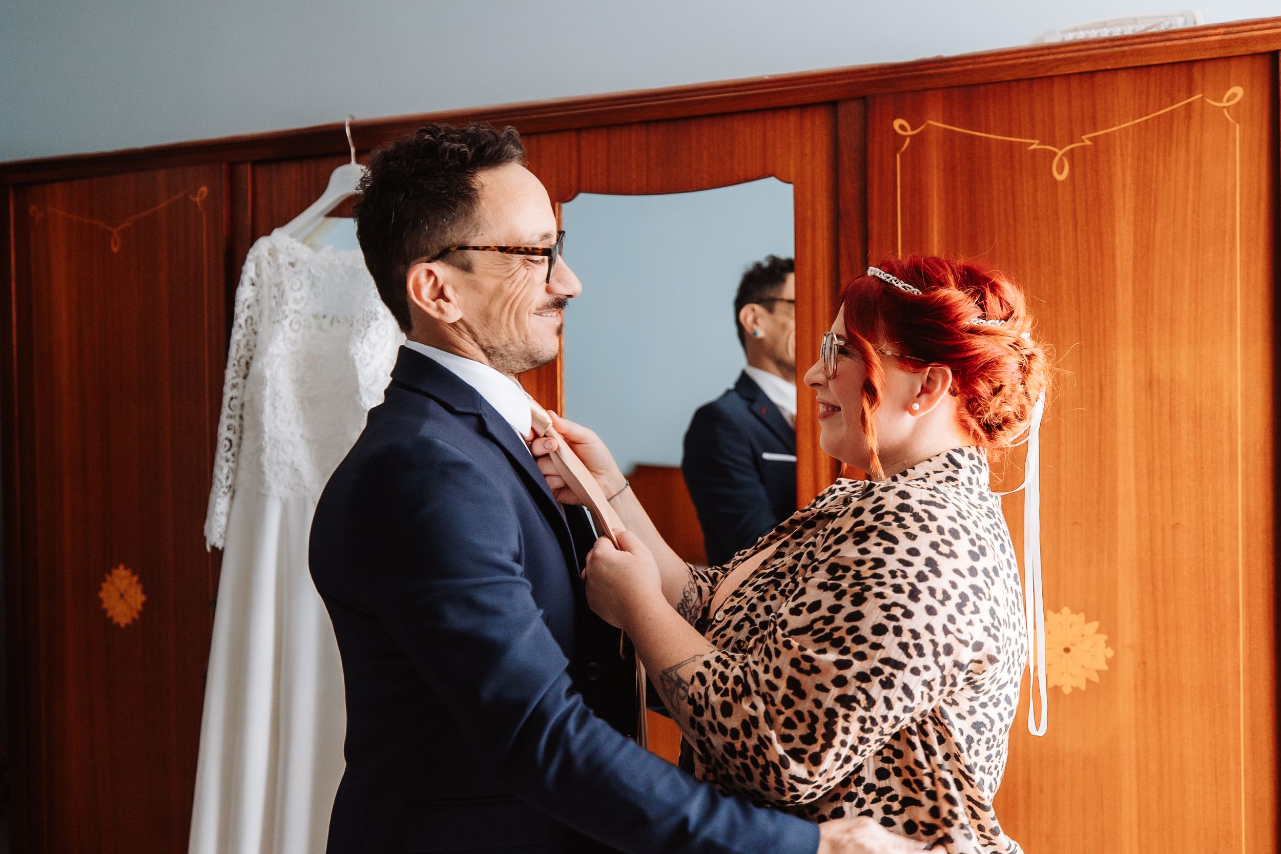 Two people stand together in a warm wood-paneled room, one adjusting the other's tie during wedding preparations.