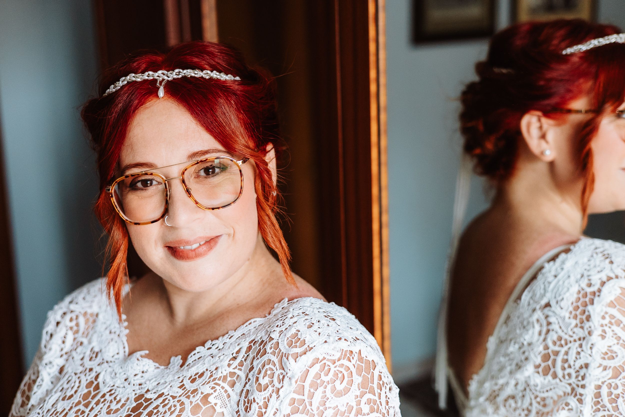 A bride wearing a white lace dress and glasses with red hair poses near a mirror at her wedding.