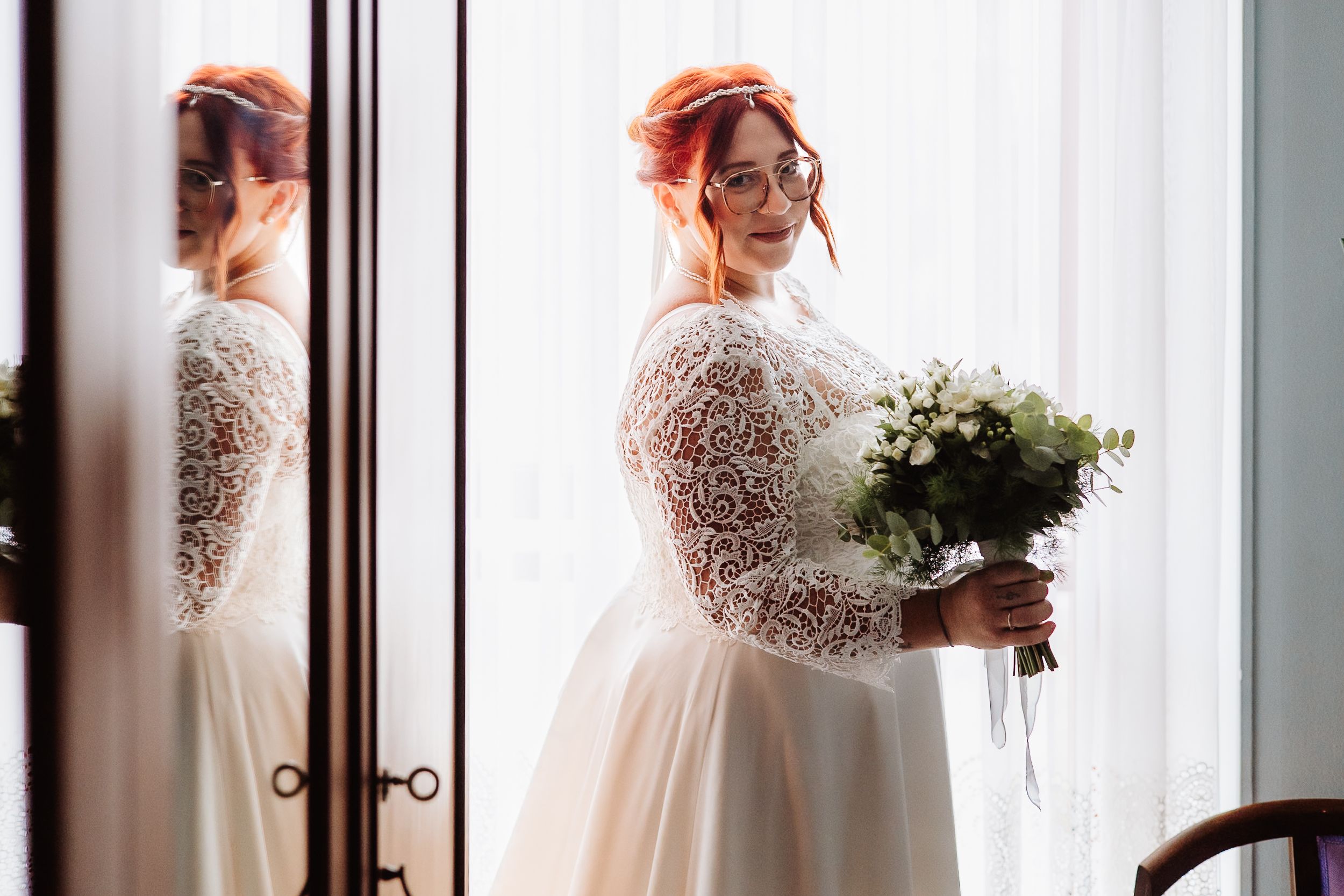 Long sleeved lace wedding dress with gathered skirt and white floral bouquet captured in natural window light.