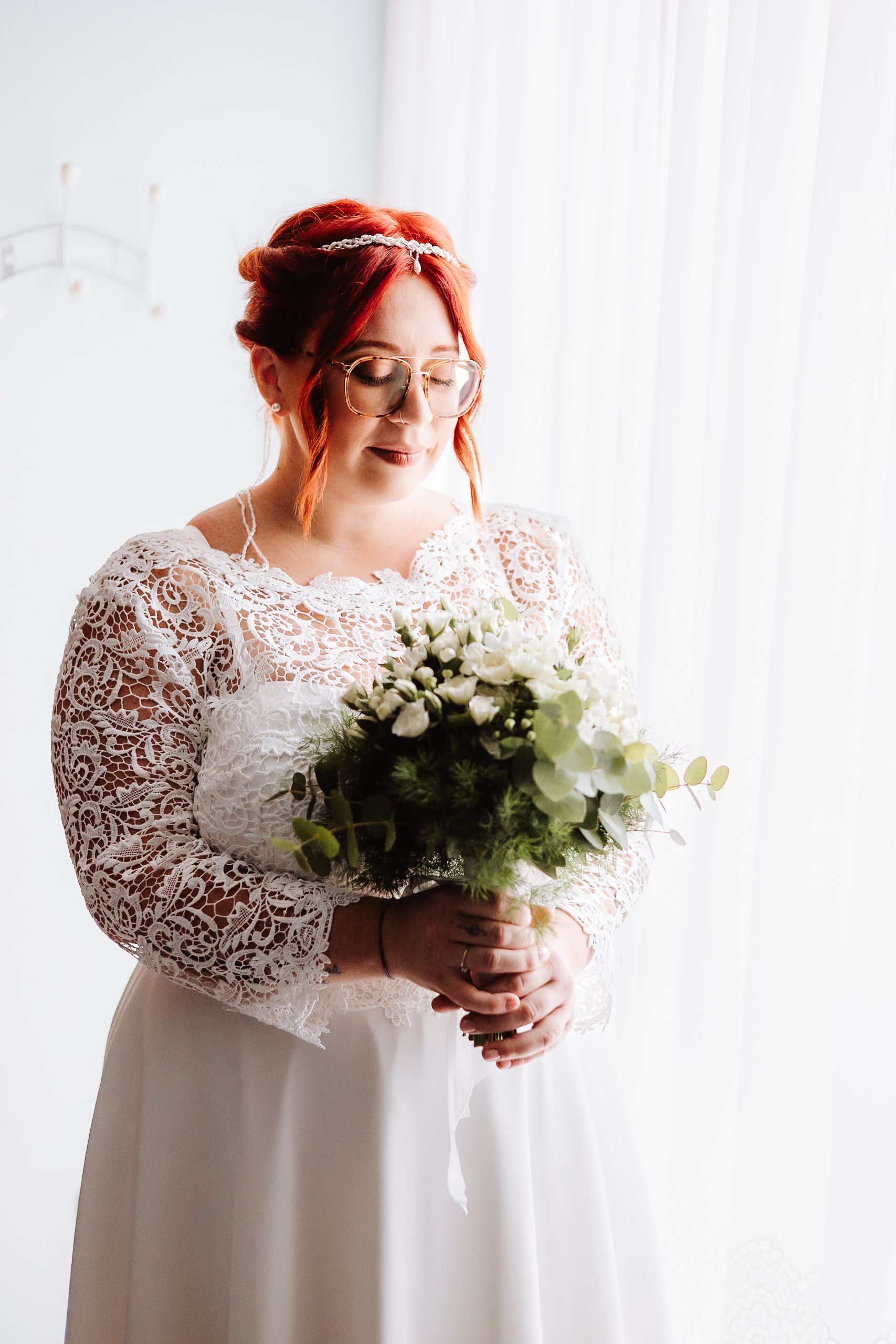 Bride in lace long sleeve wedding dress and glasses holds white floral bouquet by window in soft light.