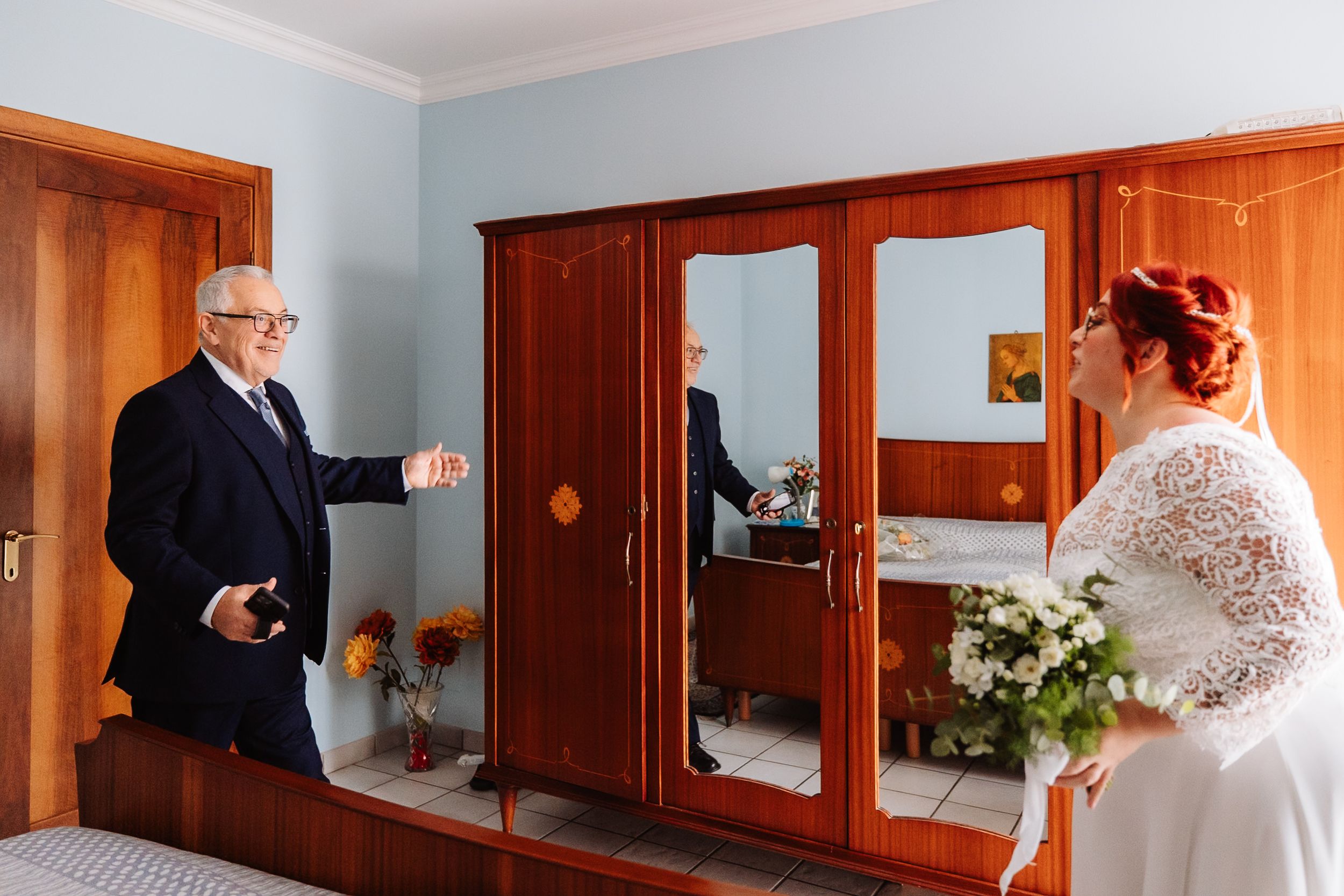 An emotional moment in a bedroom with wooden wardrobes and a mirror reflection, featuring white lace and flowers.