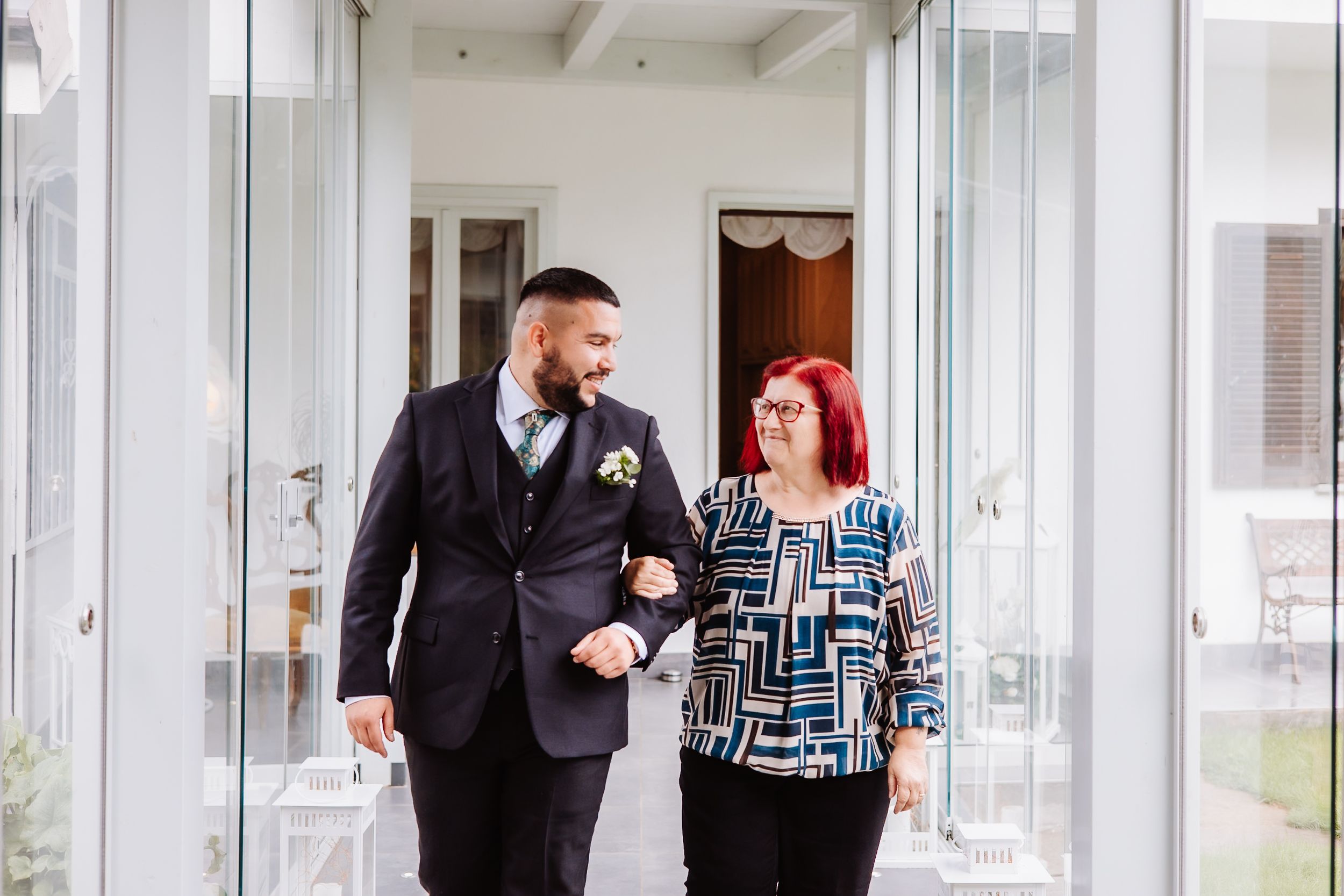 A couple link arms and walk together in formal attire along a bright white hallway.