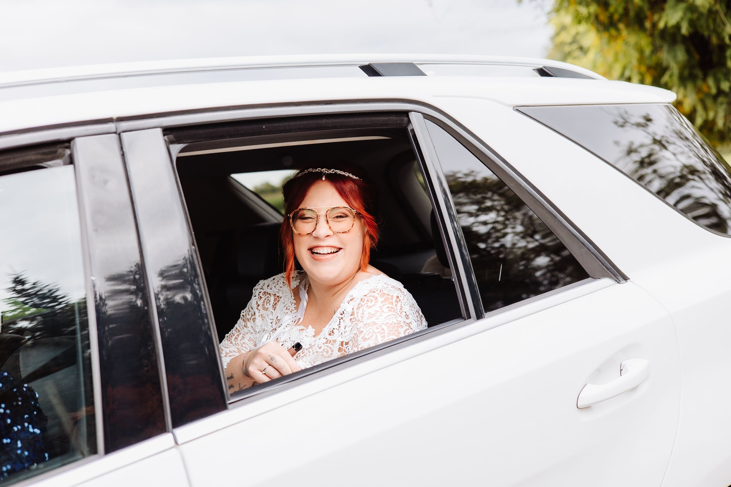 A happy passenger smiles from the back window of a white car on a sunny day.