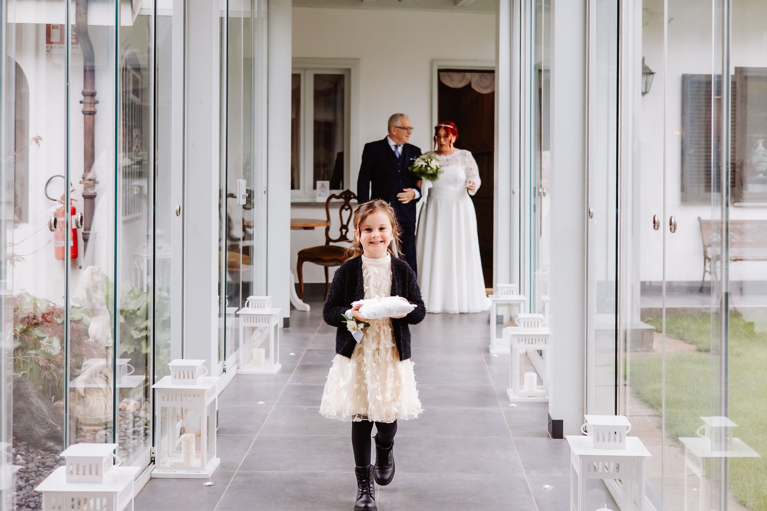A young flower girl in black boots and white dress runs joyfully down a covered porch while figures stand in doorway.