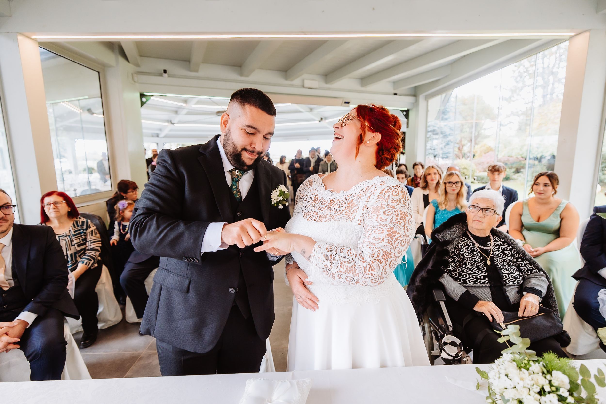 Joyful wedding couple feed each other cake at their reception while guests observe in the background.