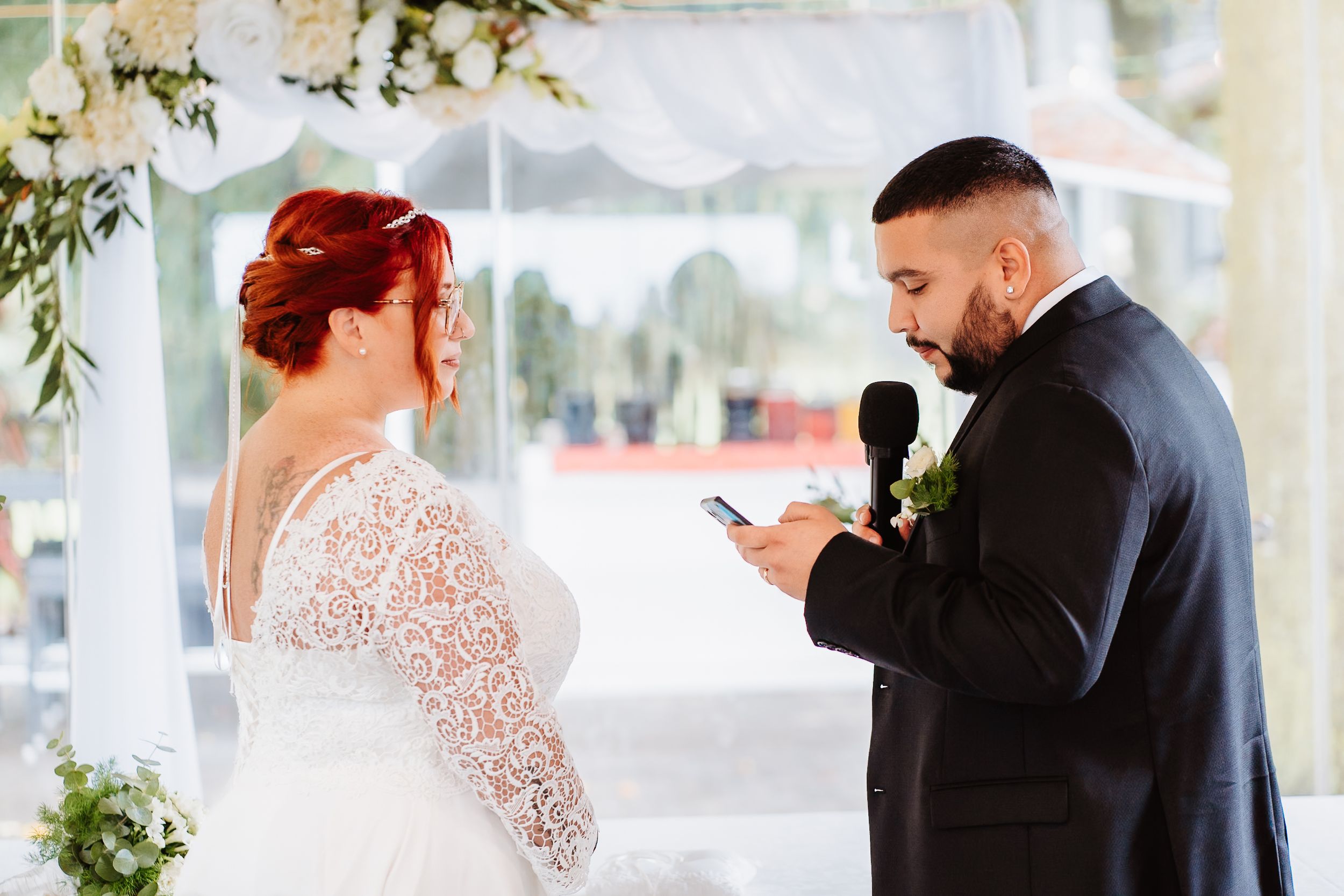 Couple exchanges wedding vows under white floral decorations at outdoor ceremony.