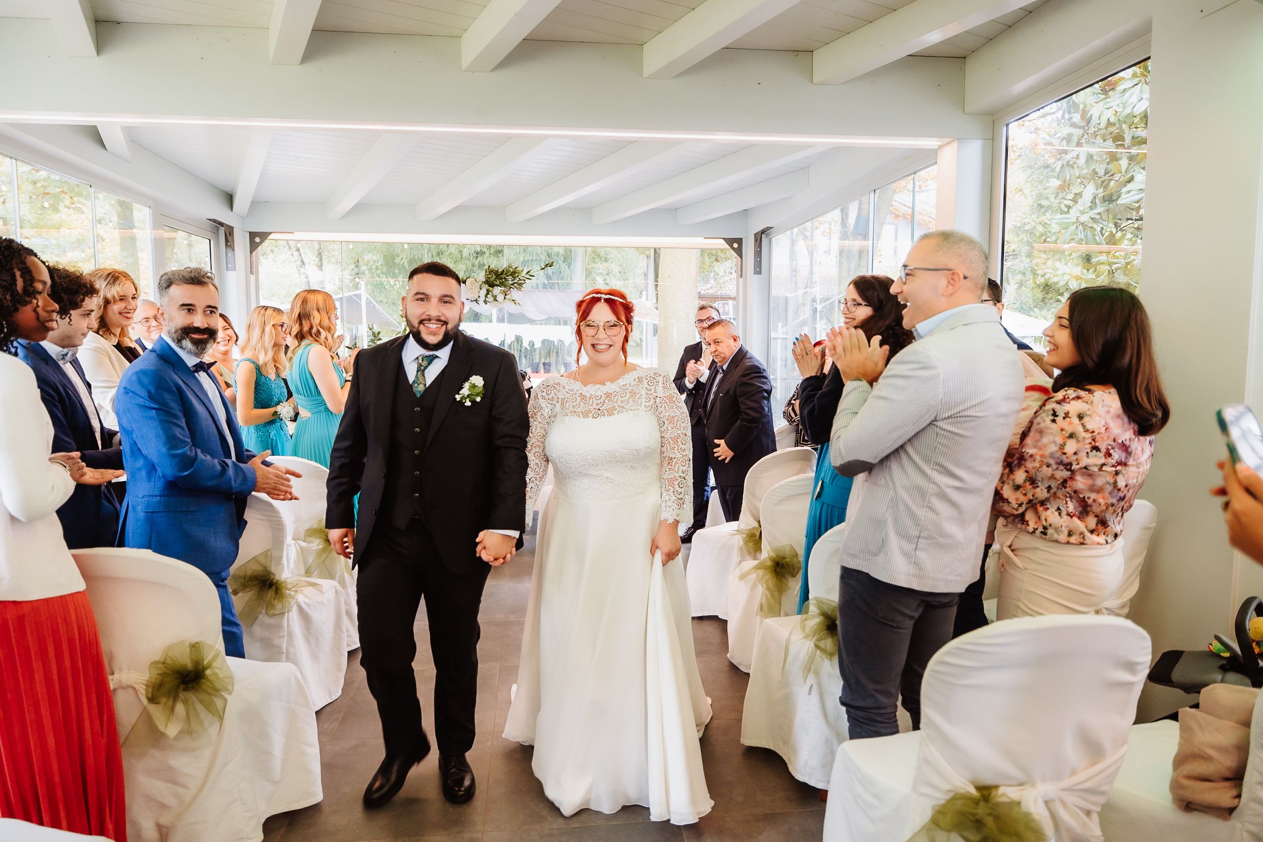 A wedding ceremony unfolds in a bright indoor venue with guests watching two people walk down an aisle between white chairs.