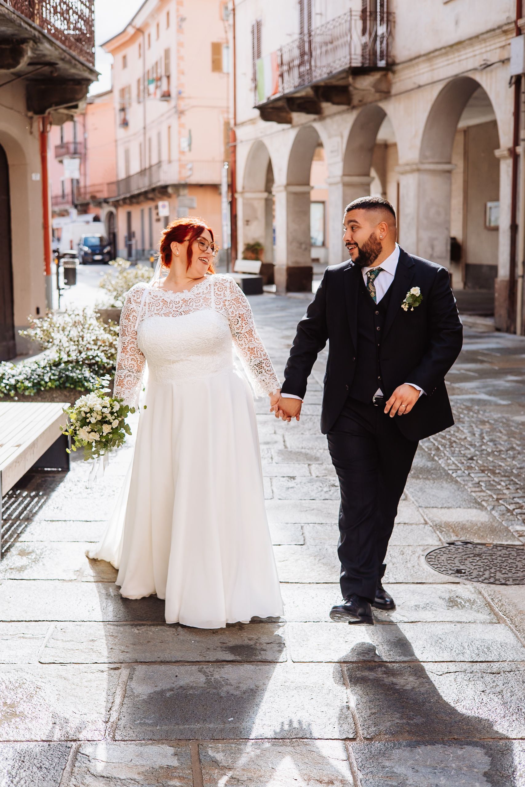 A couple in wedding attire walking hand in hand through a charming Italian street with stone arches and buildings.
