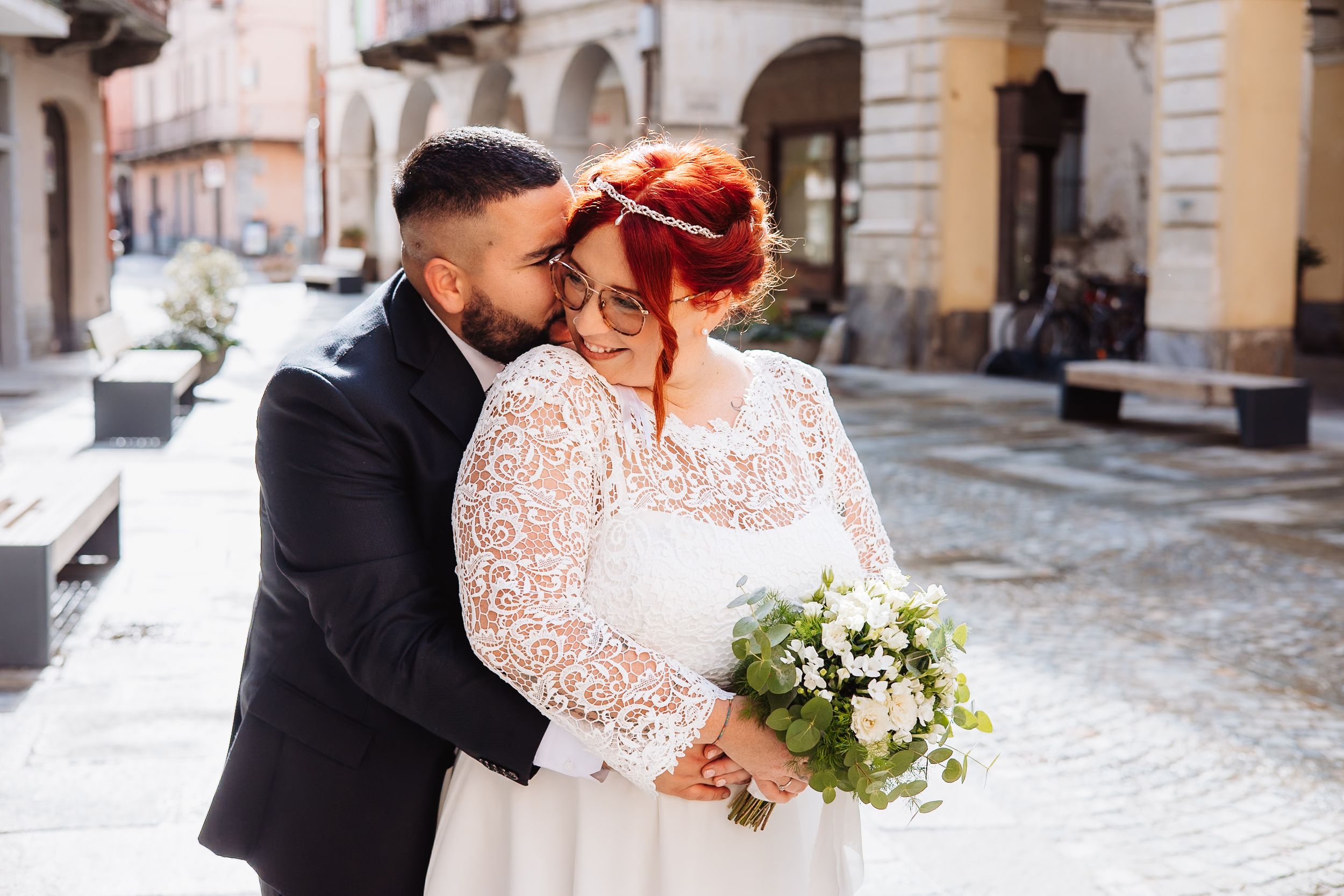 Couple in wedding attire share intimate moment in winter while holding white floral bouquet outside historic building.