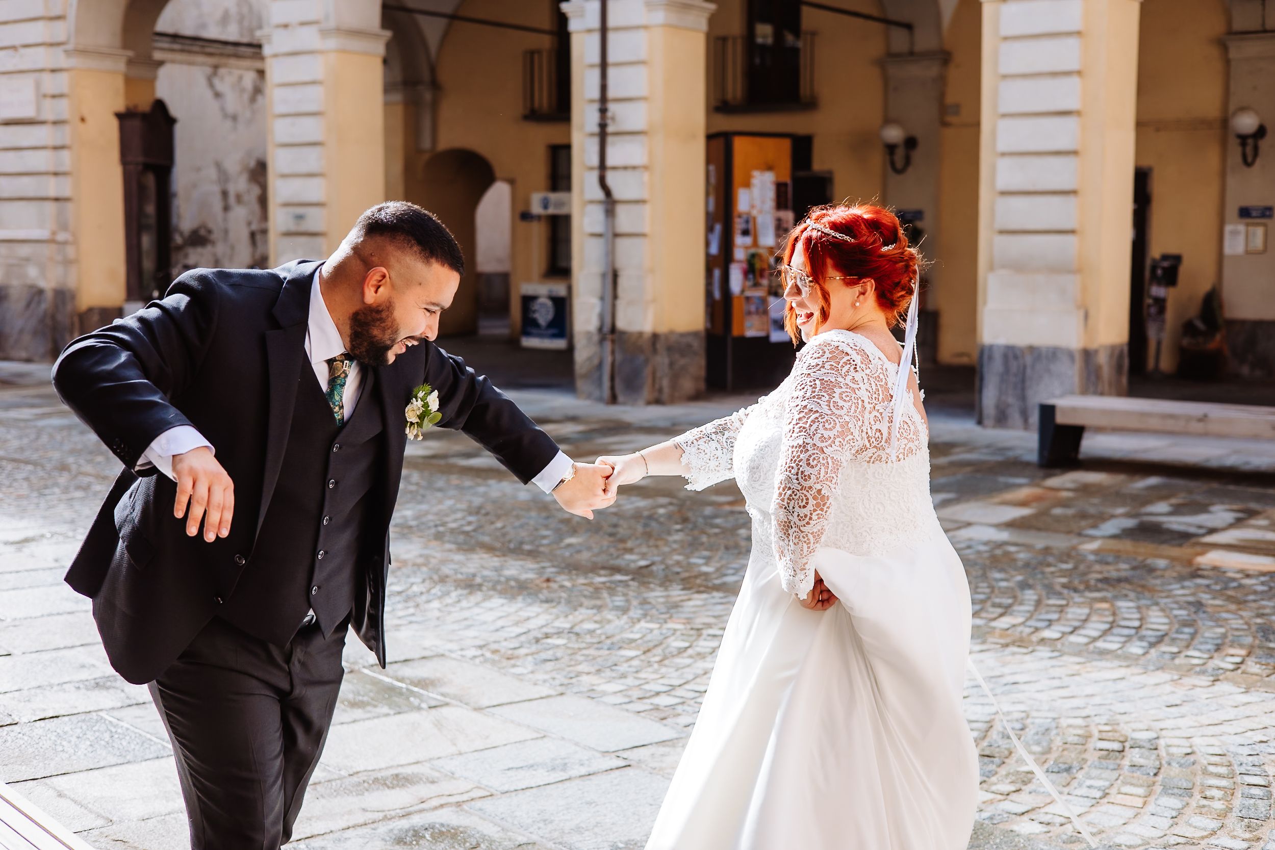 Couple in wedding attire dance playfully under arched colonnade on cobblestone street.
