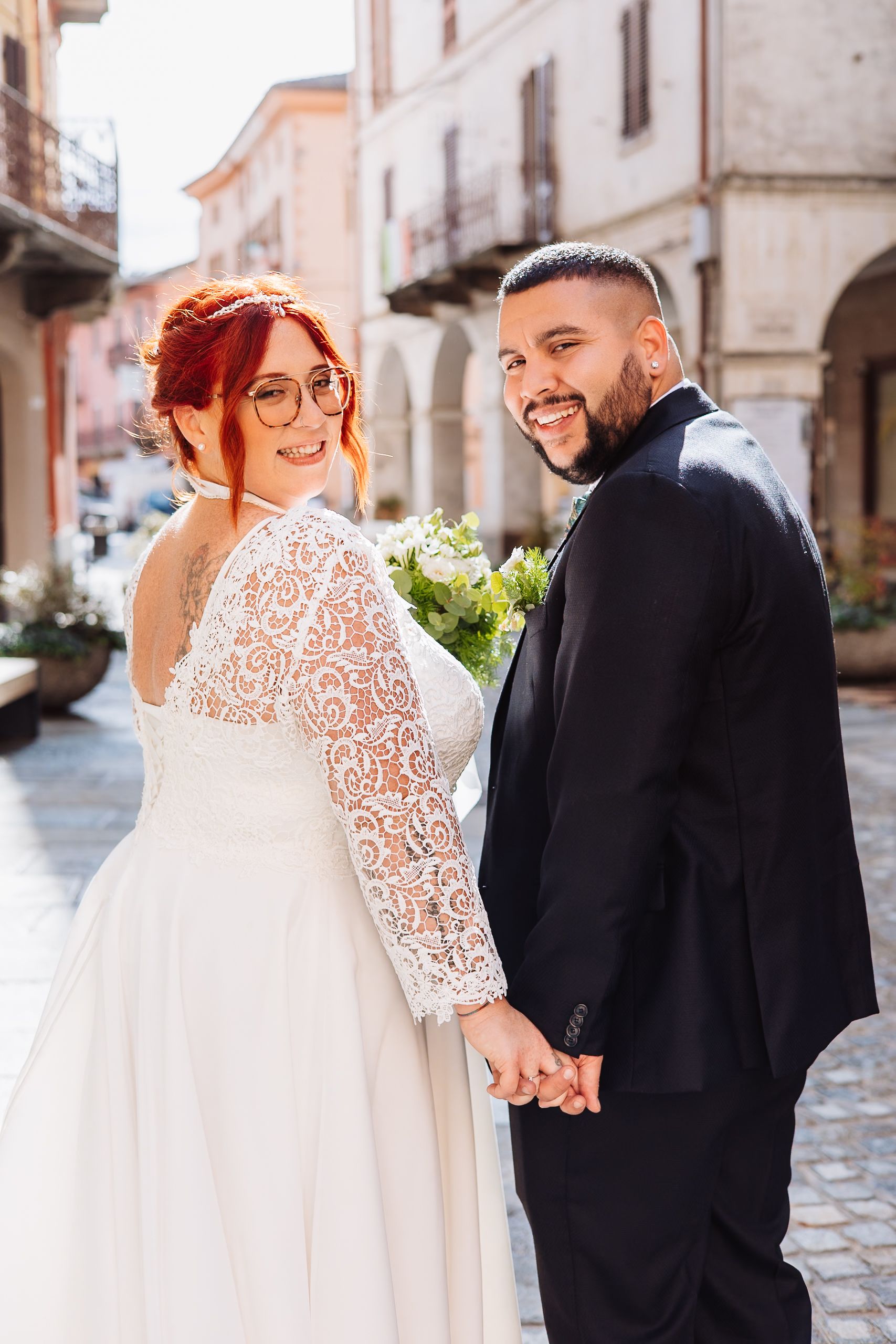 A romantic wedding photo in an Italian street with the couple holding hands, featuring intricate lace sleeves and a bouquet.