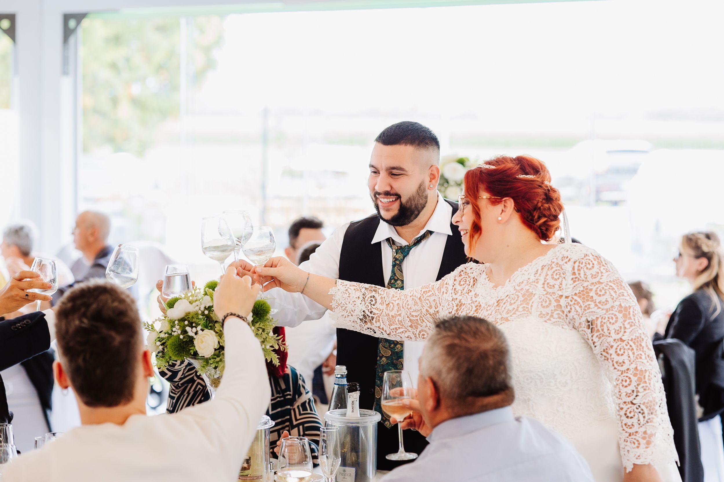 A happy couple raises their glasses in a toast during their wedding reception, surrounded by family and friends.