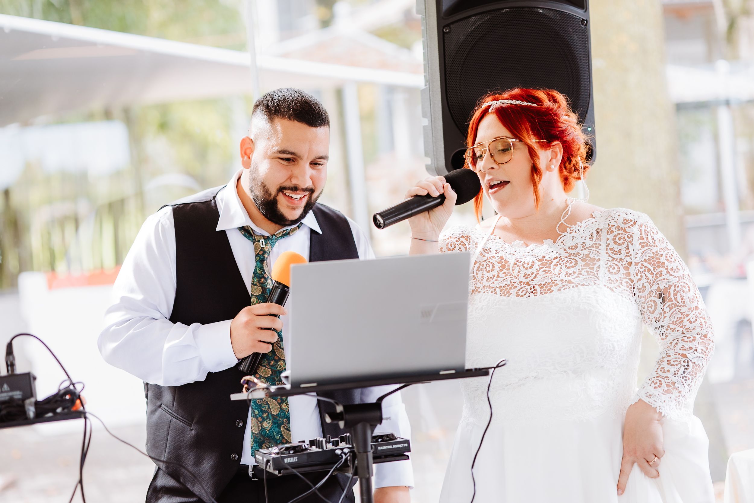 A wedding singer and singer perform together at an outdoor celebration using a laptop and microphones.