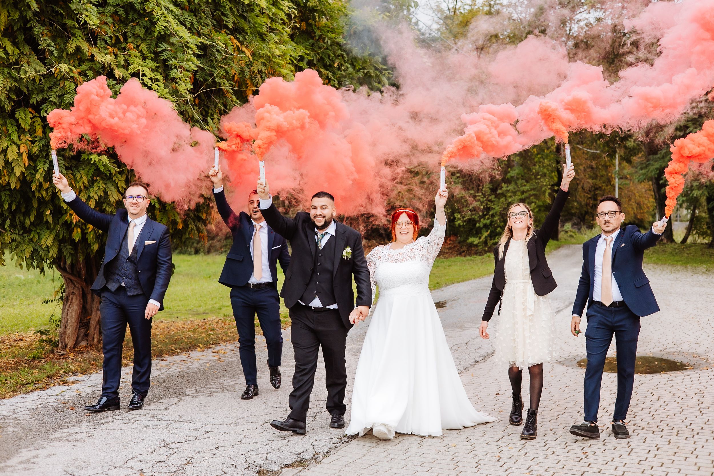 A wedding party celebrates outdoors with pink smoke bombs creating dramatic colorful clouds on a path near trees.