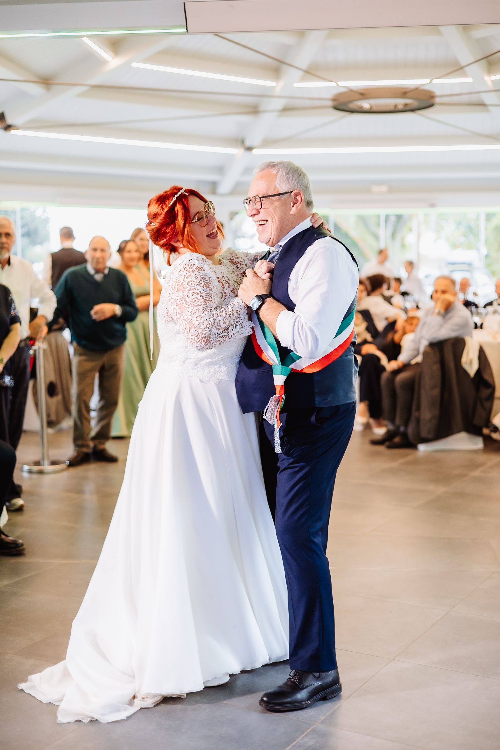 A couple shares their first dance at a wedding reception in a bright indoor venue with white decor and guest tables.