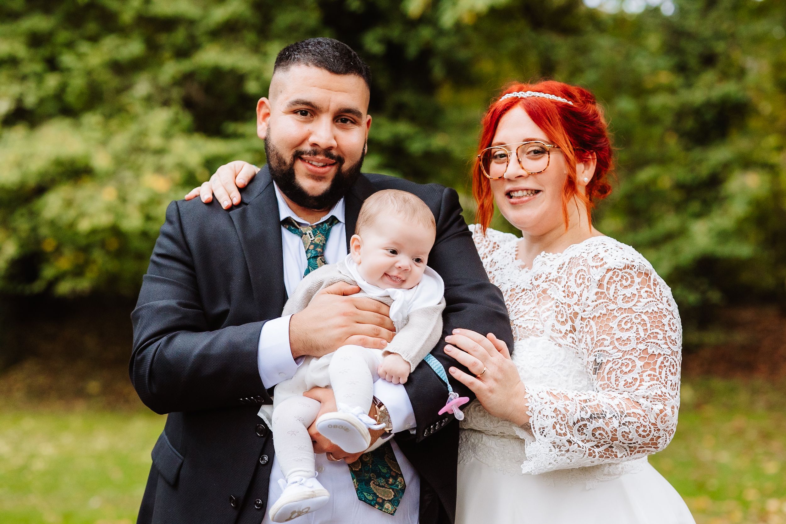 A couple in wedding attire stands outdoors holding a baby in white, with green foliage in the background.