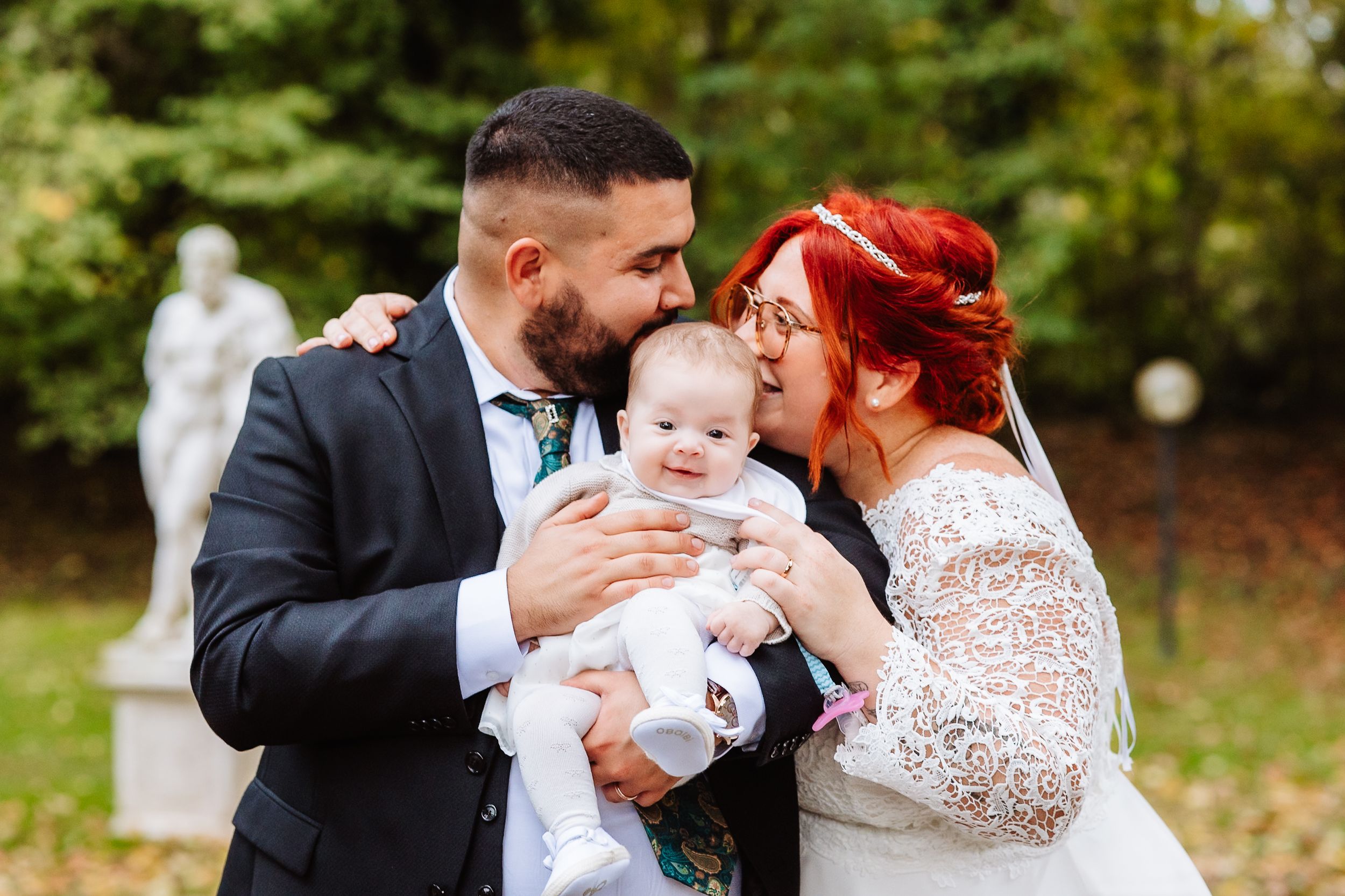 A couple in wedding attire share a tender moment with their baby outside during an autumn day.