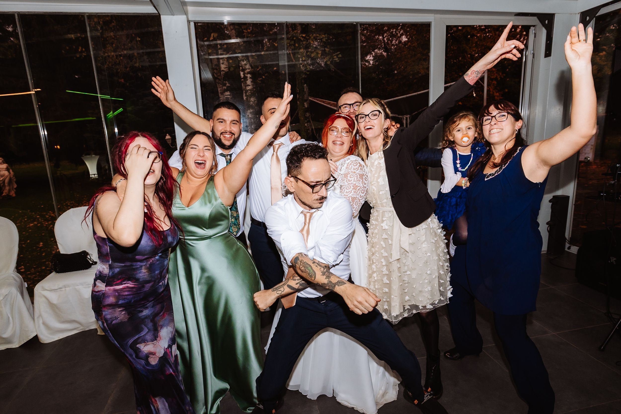 A group of wedding guests dancing enthusiastically at a reception venue with large windows in the background.