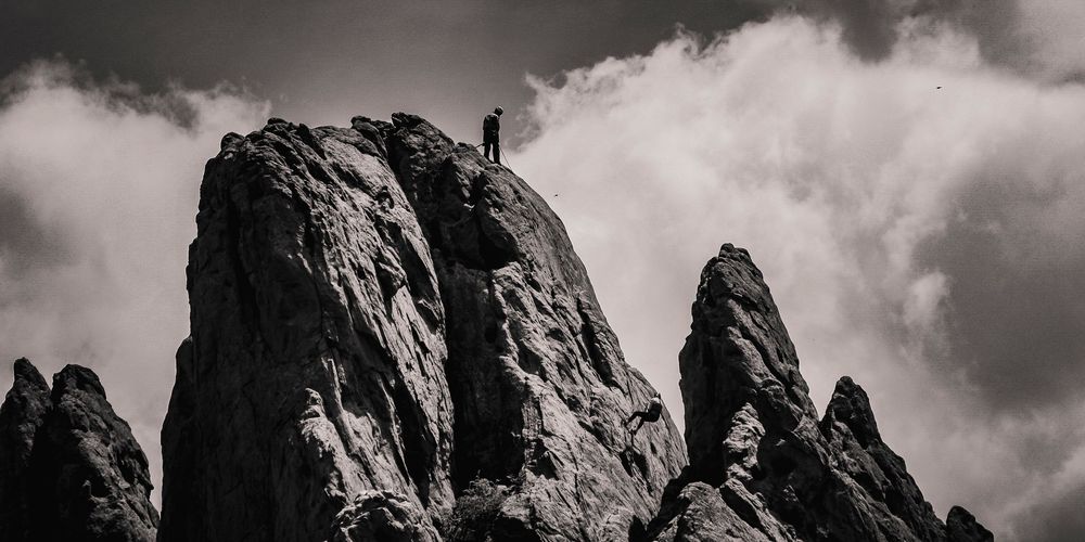 Black and white view of a climber silhouetted atop a dramatic rocky mountain peak against cloudy skies.
