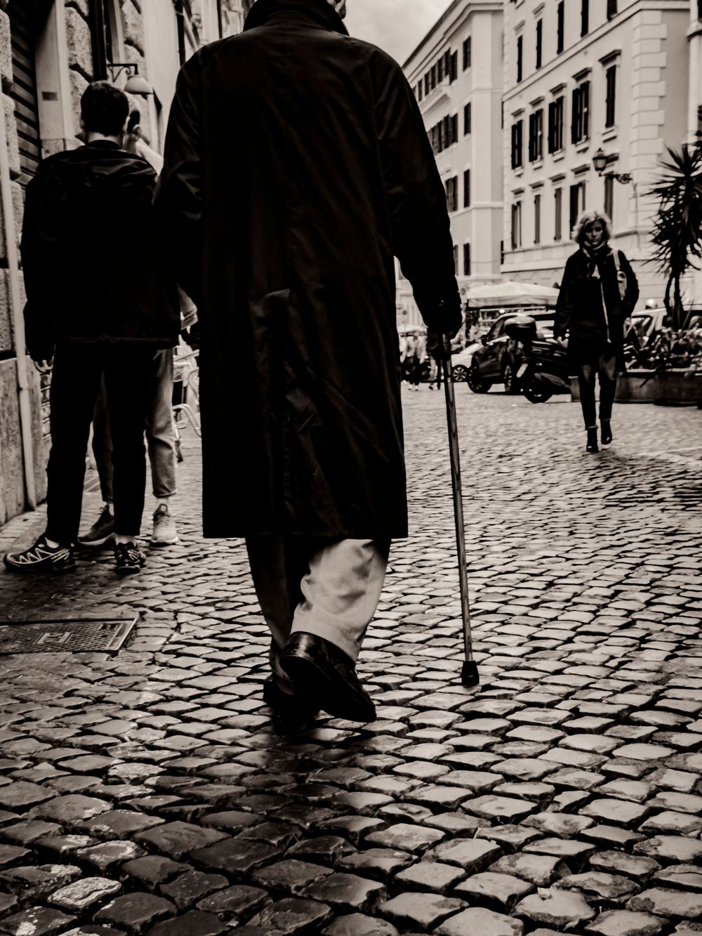 Black and white street photo showing person walking with cane on cobblestone street in European city.