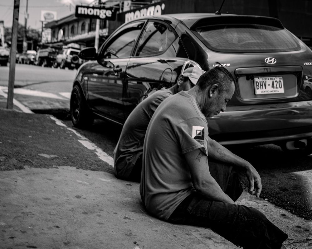 Urban street scene in black and white showing someone seated on a curb next to parked cars.