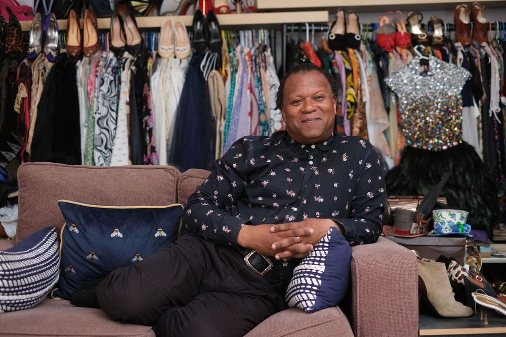 Someone sits comfortably on a brown couch with a colorful wardrobe and clothing rack visible in the background.
